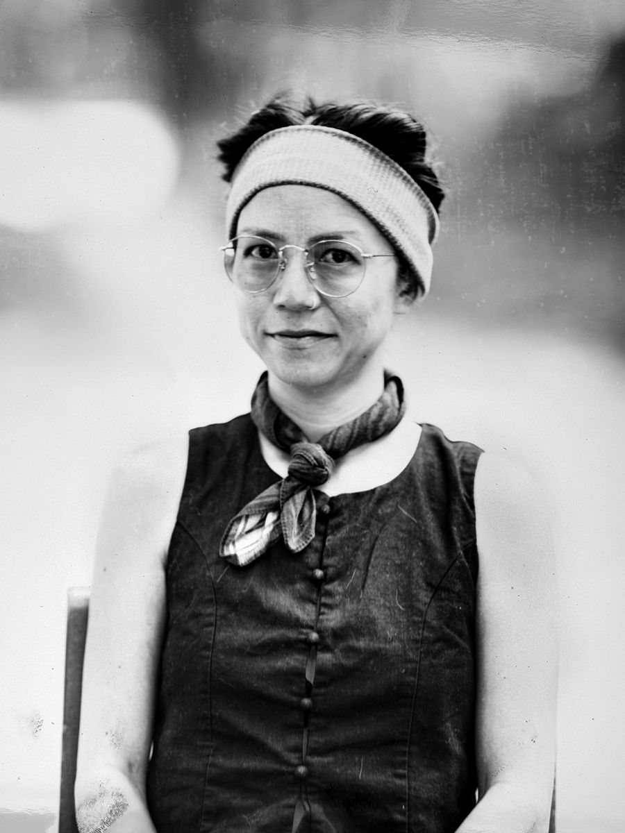 Portrait of a woman in a headband, glasses, and a dark vest with a knotted scarf.